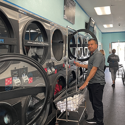 Laundromat in San Diego & Chula Vista - Close-up of a person holding a white plastic hamper filled with laundry in front of laundromat washing machines
